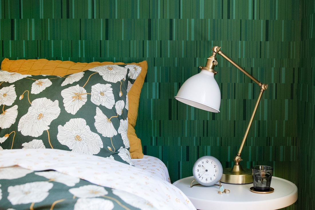 Bed with green floral duvet and mustard pillow next to brass lamp and round alarm clock against green striped wallpaper