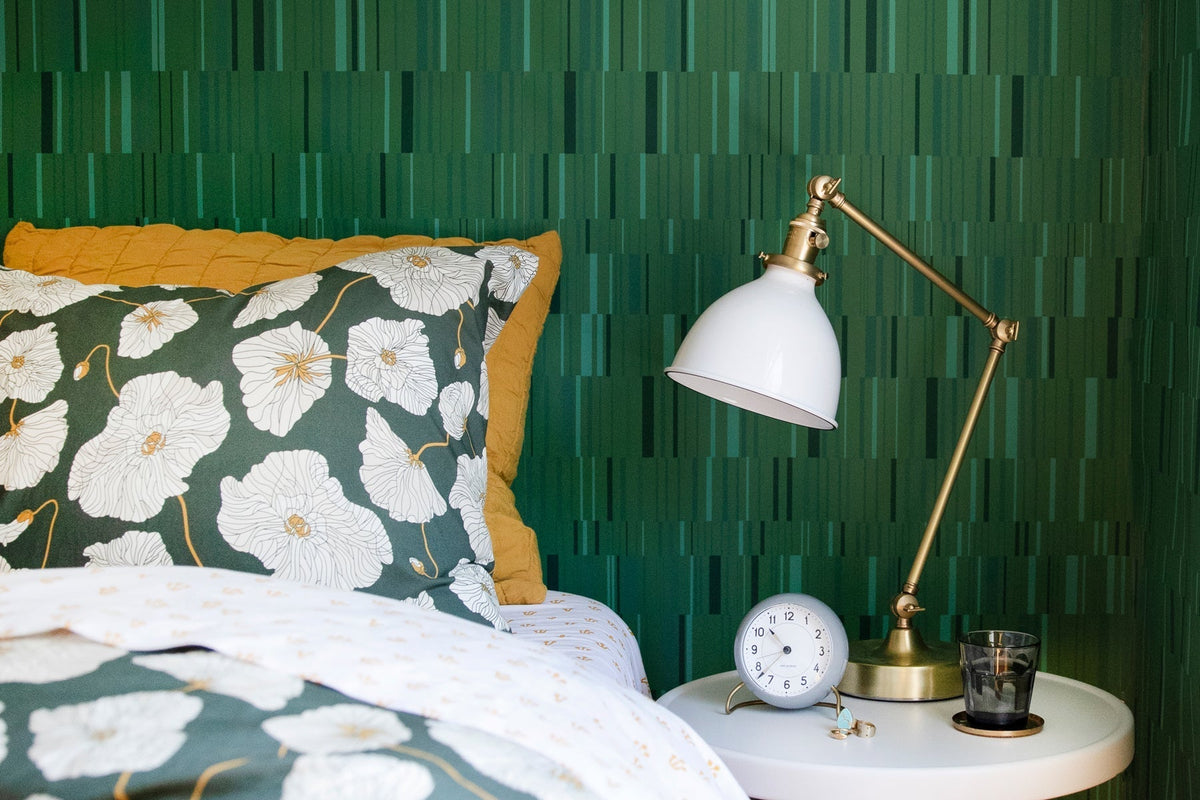 Bed with green floral duvet and mustard pillow next to brass lamp and round alarm clock against green striped wallpaper