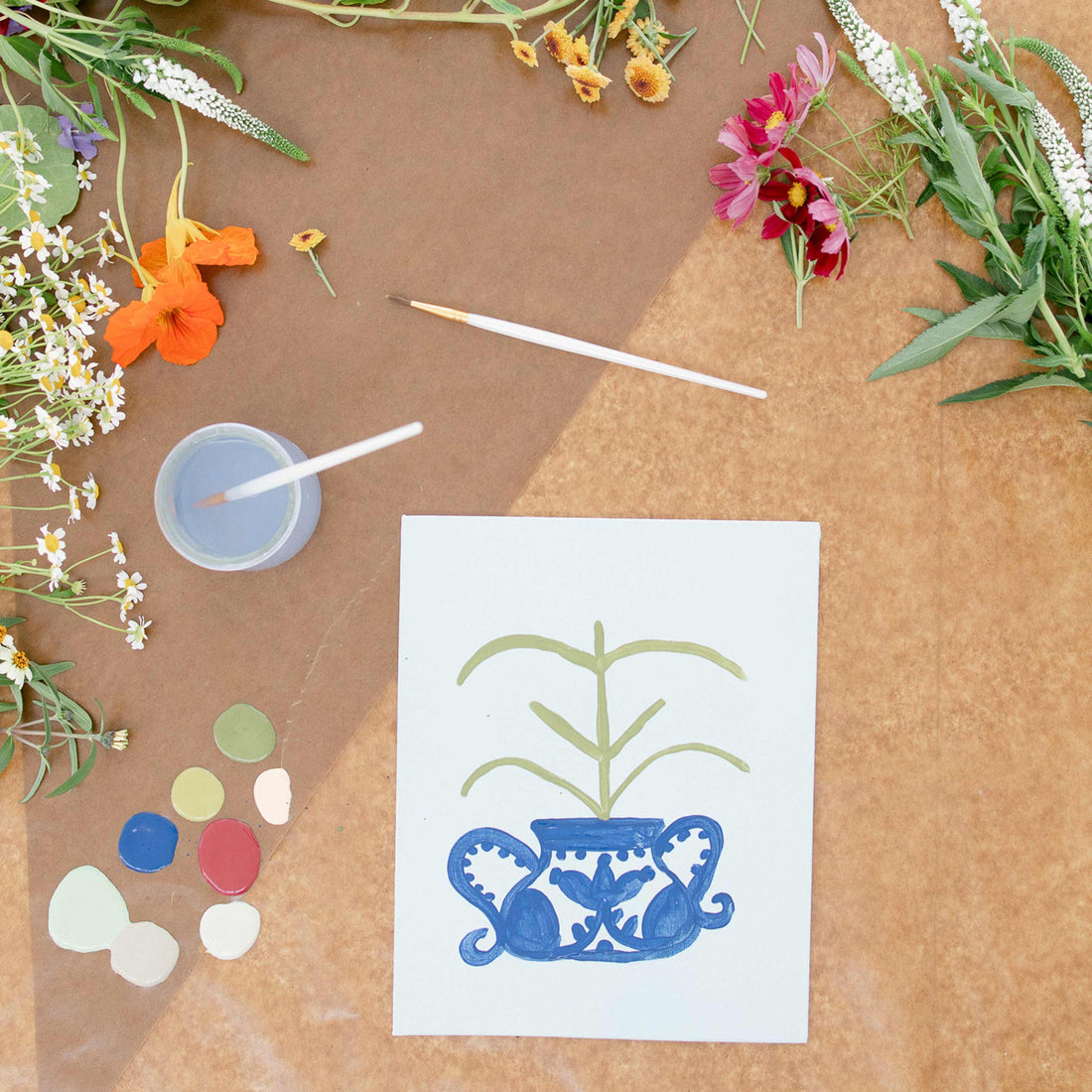 Overhead view of a craft workspace with flowers, paintbrushes, a paint cup and a hand-painted botanical on paper