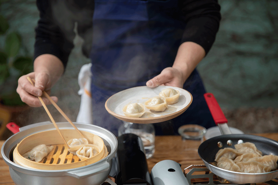 Person holding a plate of steamed pork and chive dumplings while using chopsticks to lift dumplings from a bamboo steamer
