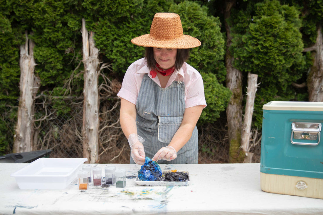 Person in a straw hat and striped apron ice-dyeing a bundled piece of fabric on an outdoor table with trays and dye powders