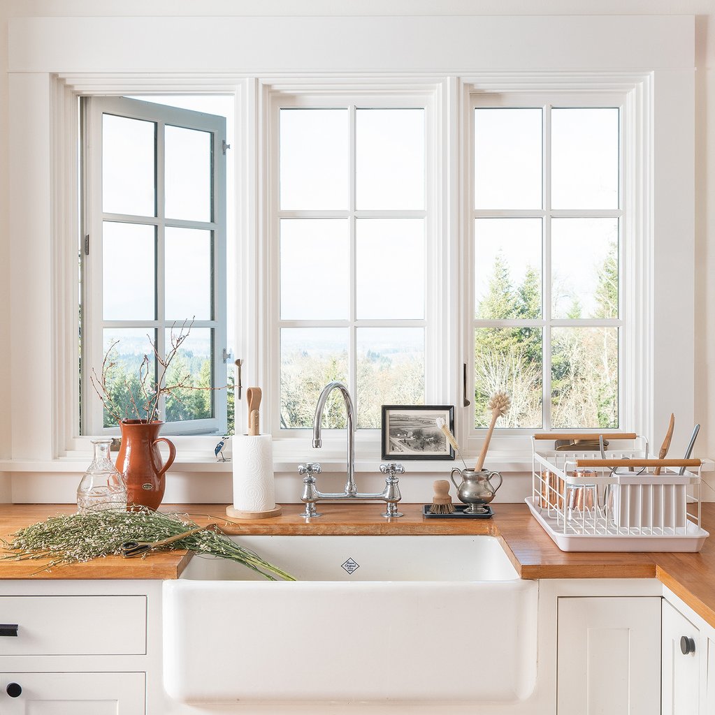 Farmhouse sink with chrome faucet, wooden countertops, drying rack and paper towel under sunlit casement windows overlooking trees
