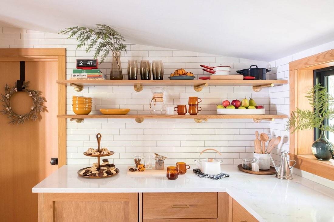 Holiday cutout cookies on a two-tier stand on a kitchen counter with a white teapot, amber glass mugs, open wooden shelves with bowls and a bowl of fruit, and a fern by the window