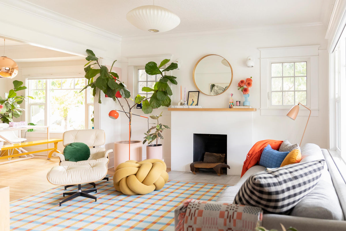 Bright family living room styled with an Eames-style lounge chair, yellow knot ottoman, checkered rug, fiddle-leaf fig, round mirror, and colorful pillows