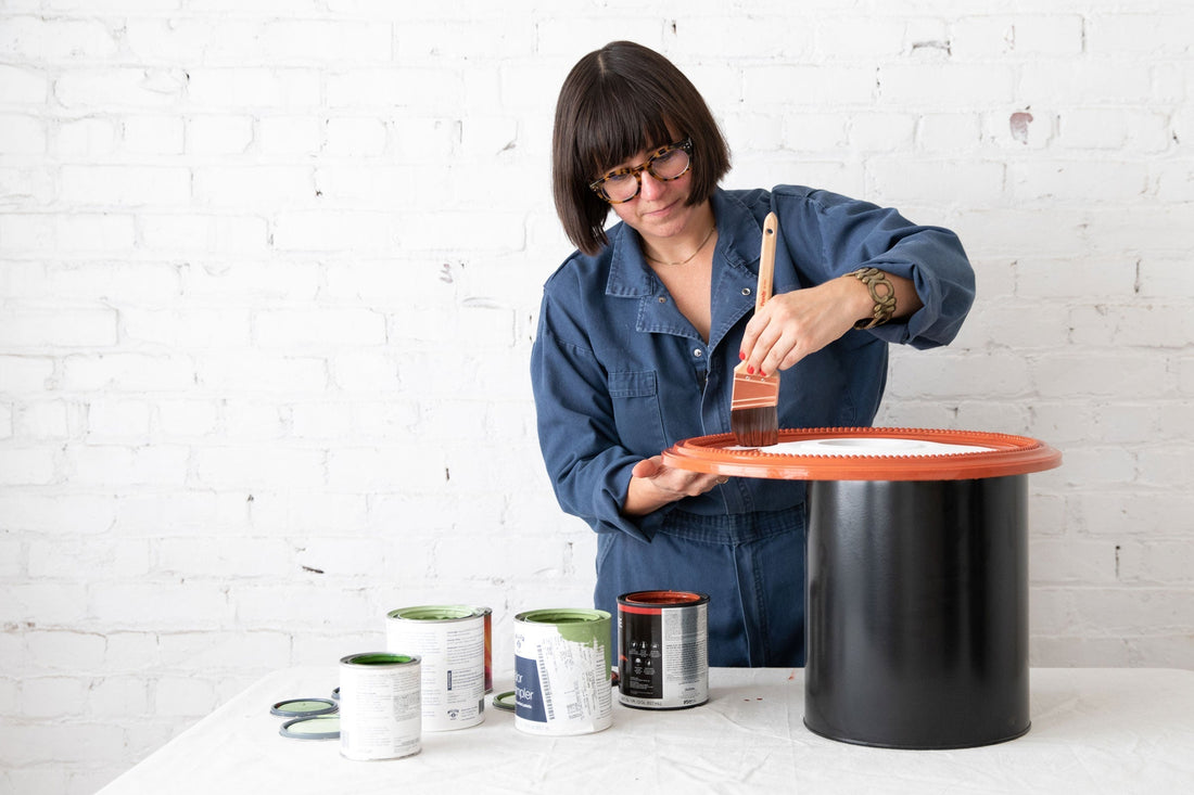 Woman painting the front of a round ceiling medallion orange at a table with open paint cans, white brick wall background