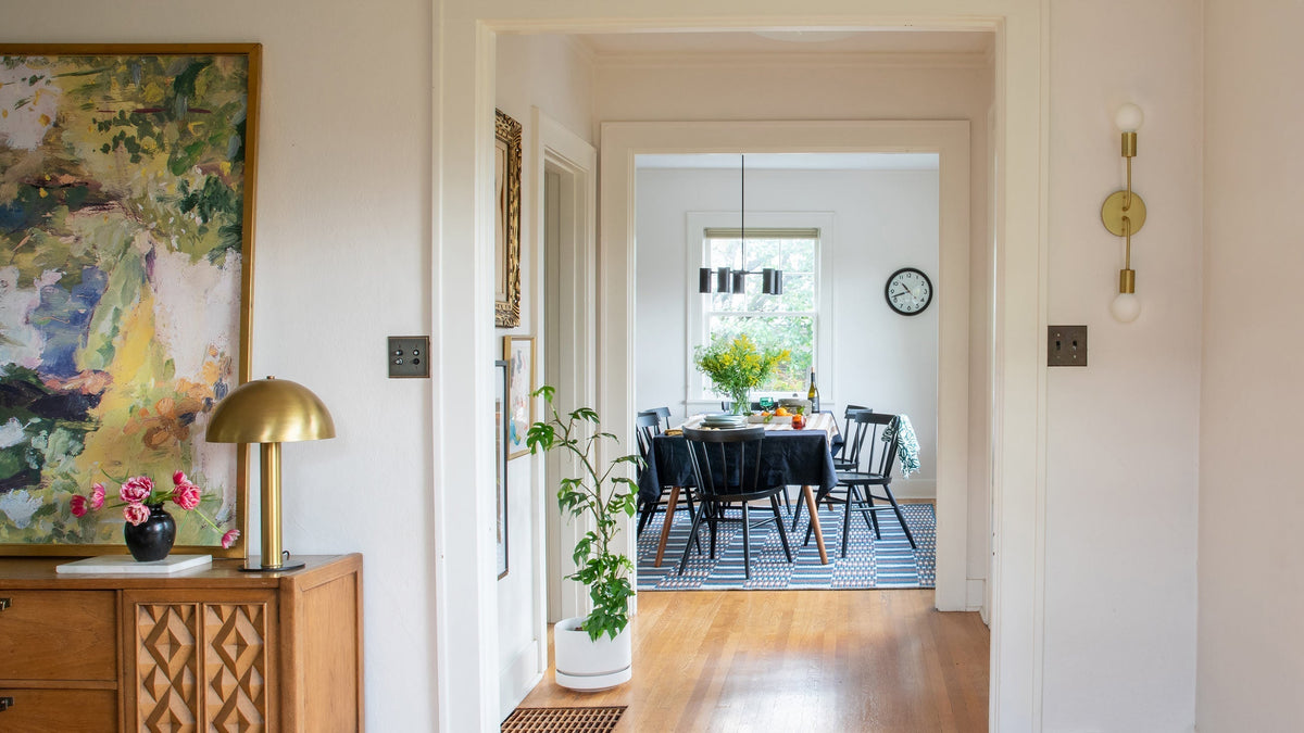 Hallway view into a bright dining room with a black table and chairs, patterned rug, yellow flower centerpiece, and brass lamp