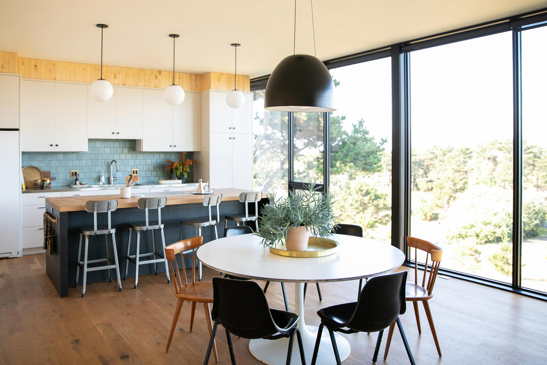 Sunlit kitchen and dining area with round white table, potted plant, wood island with metal stools, and large windows