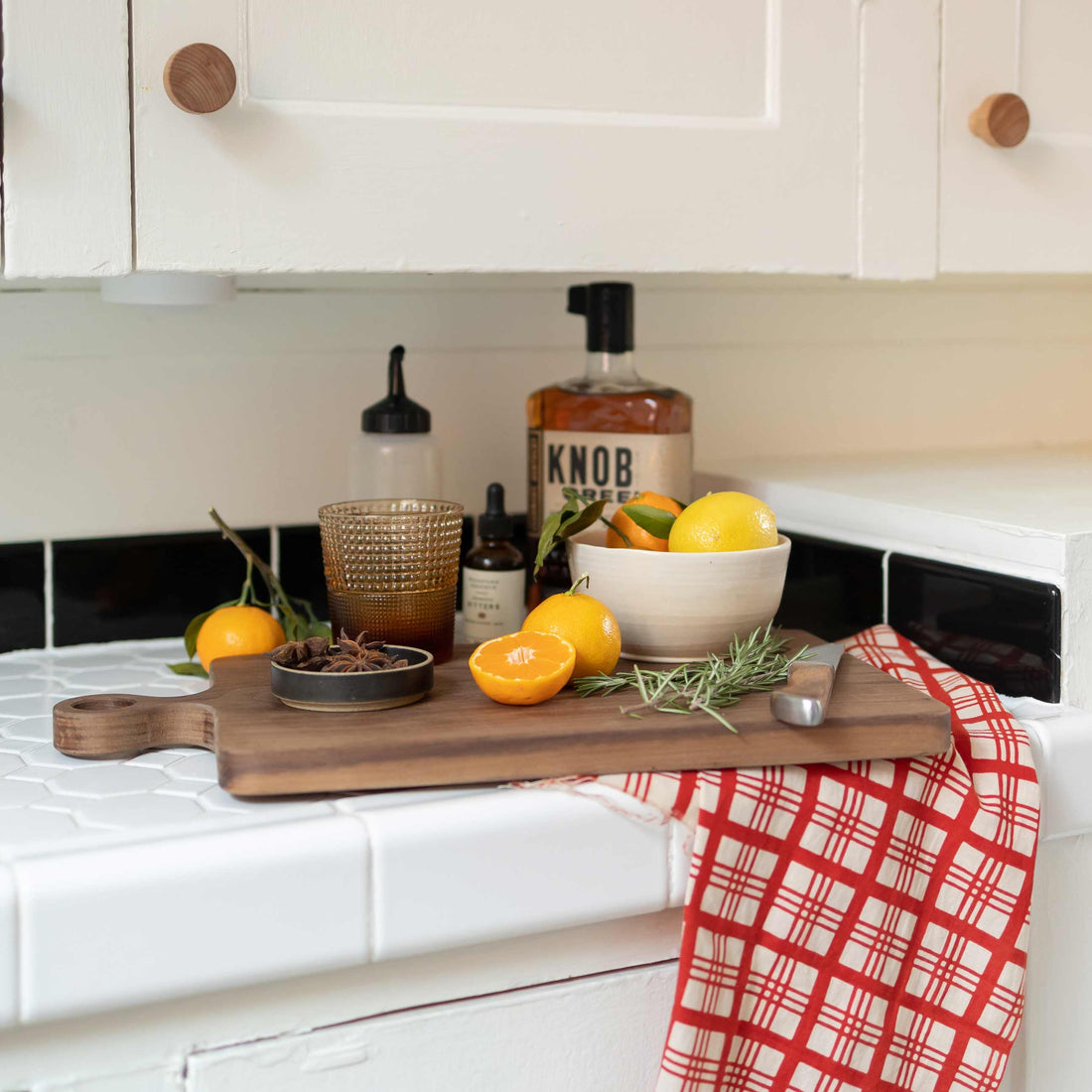 Wood cutting board with oranges and a halved clementine, star anise and rosemary, glass and a whiskey bottle on a white tiled kitchen counter