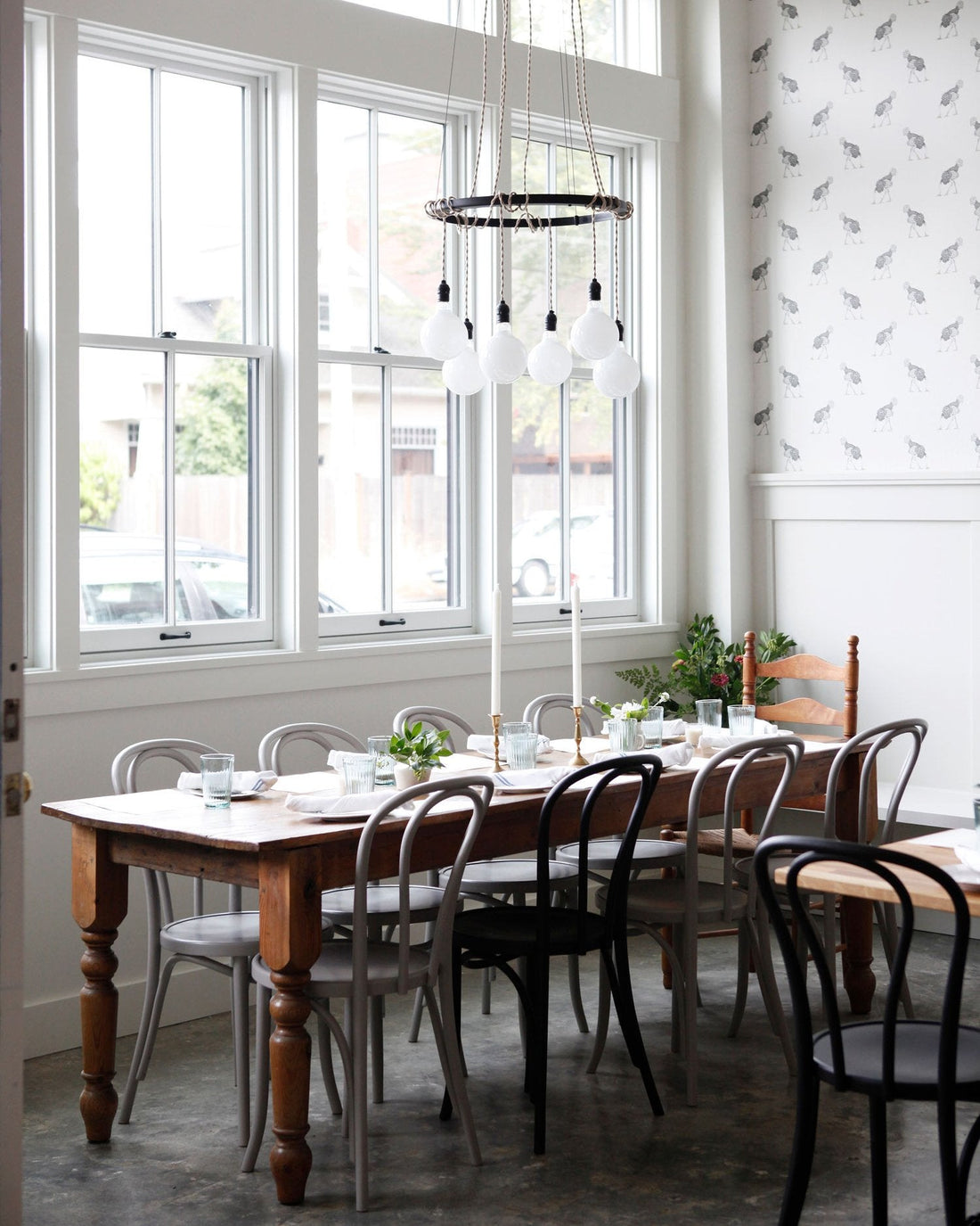 Dining table set with glassware beneath a rope-wrapped ring chandelier in a bright restaurant dining room with tall windows