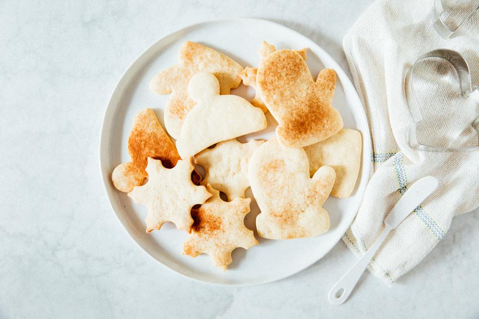 Plate of mitten- and snowflake-shaped baked cookies arranged on a marble counter with metal cookie cutters and a folded tea towel