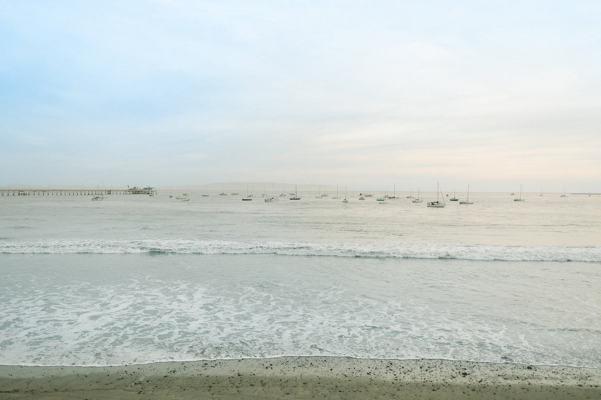 Sailboats moored offshore with a long pier to the left and gentle waves under a pale morning sky