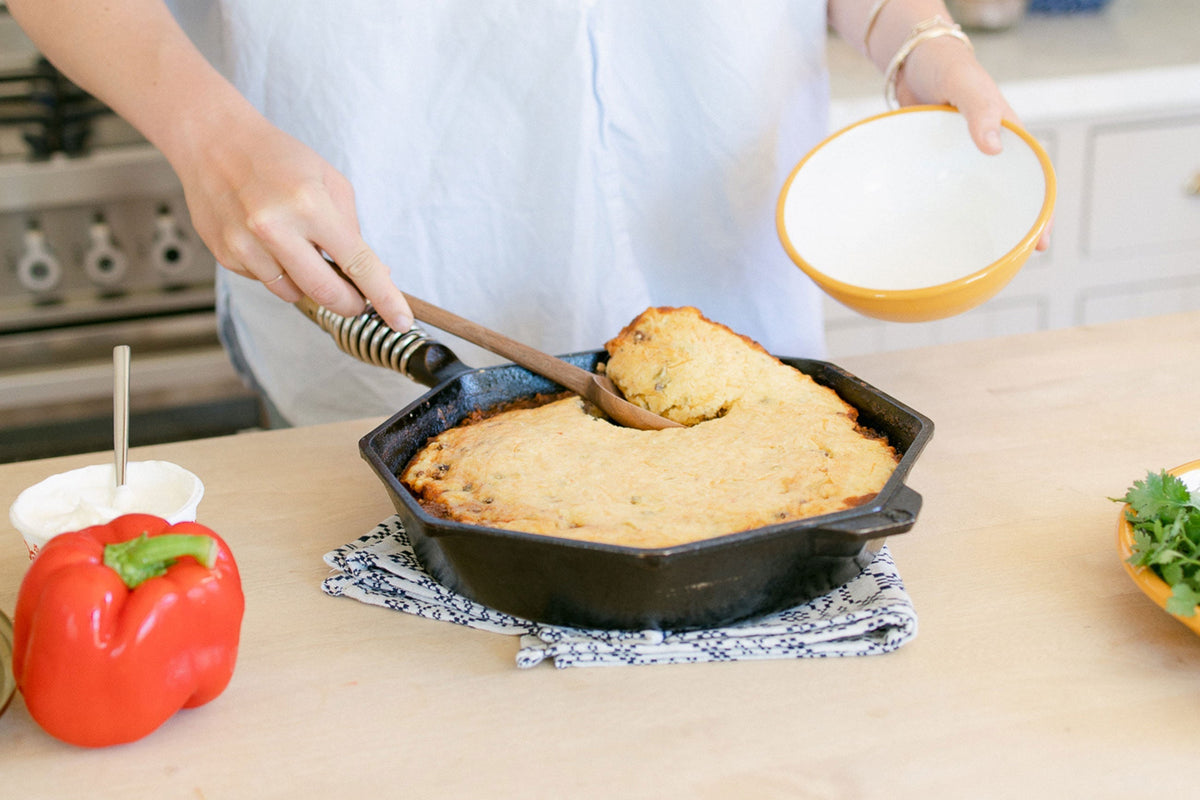 Person scooping a slice of tamale pie from a cast-iron skillet on a kitchen counter