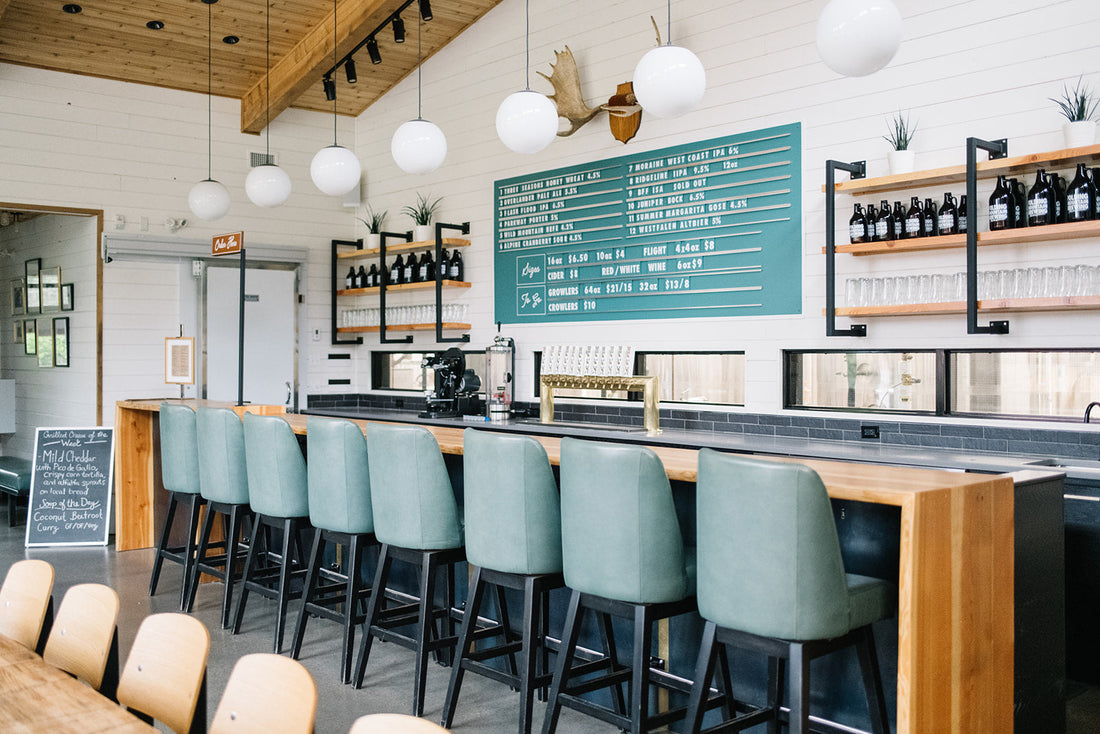 Row of teal bar stools along a wooden bar in a bright cafe interior with globe pendant lights and a teal menu board