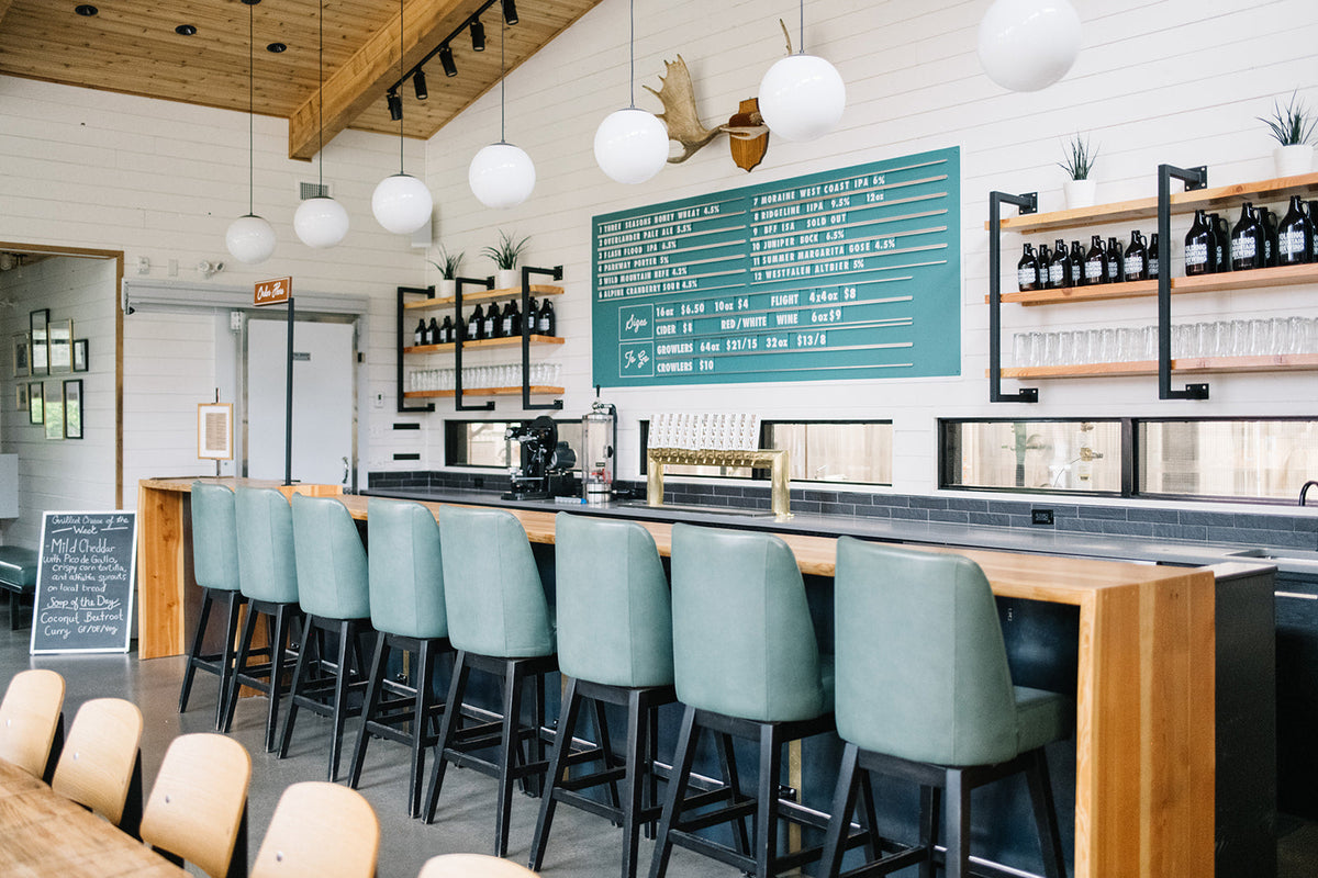 Row of teal bar stools along a wooden bar in a bright cafe interior with globe pendant lights and a teal menu board