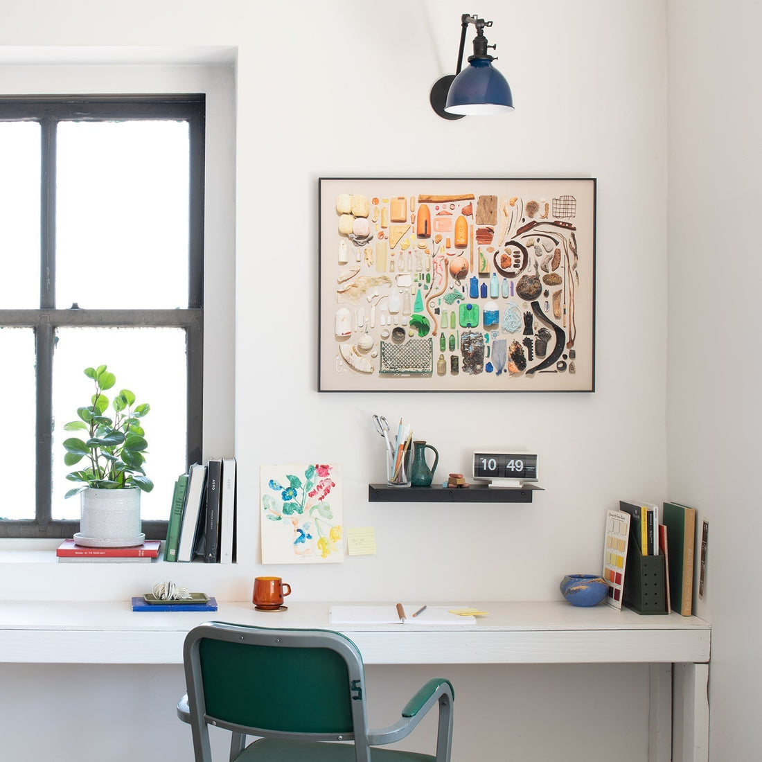 White home desk with framed Beachcomber print, green chair, window with potted plant, wall shelf with pens and flip clock reading 10:49