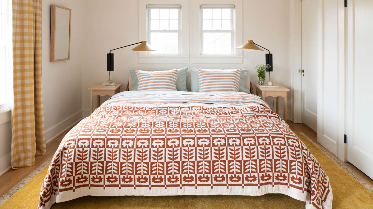 Bed with rust-colored floral quilt and striped pillows in a sunlit minimalist bedroom with yellow rug and brass swing-arm lamps