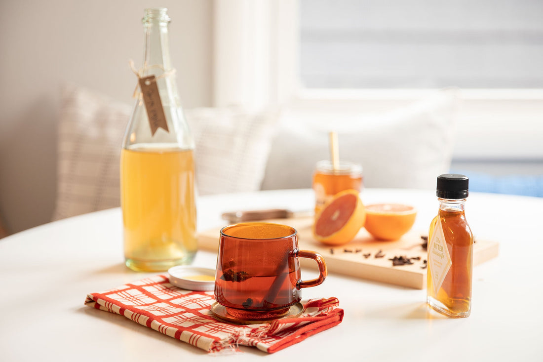 Amber mug of mulled pear brandy on a red napkin, with pear brandy bottle, bitters and halved orange on a sunlit table