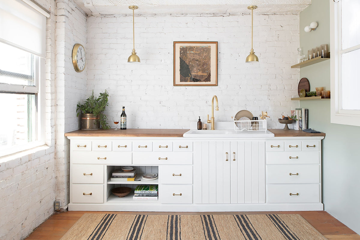 Kitchen sink area with brass faucet and two brass pendant lights, white cabinets and wood countertop against a painted white brick wall