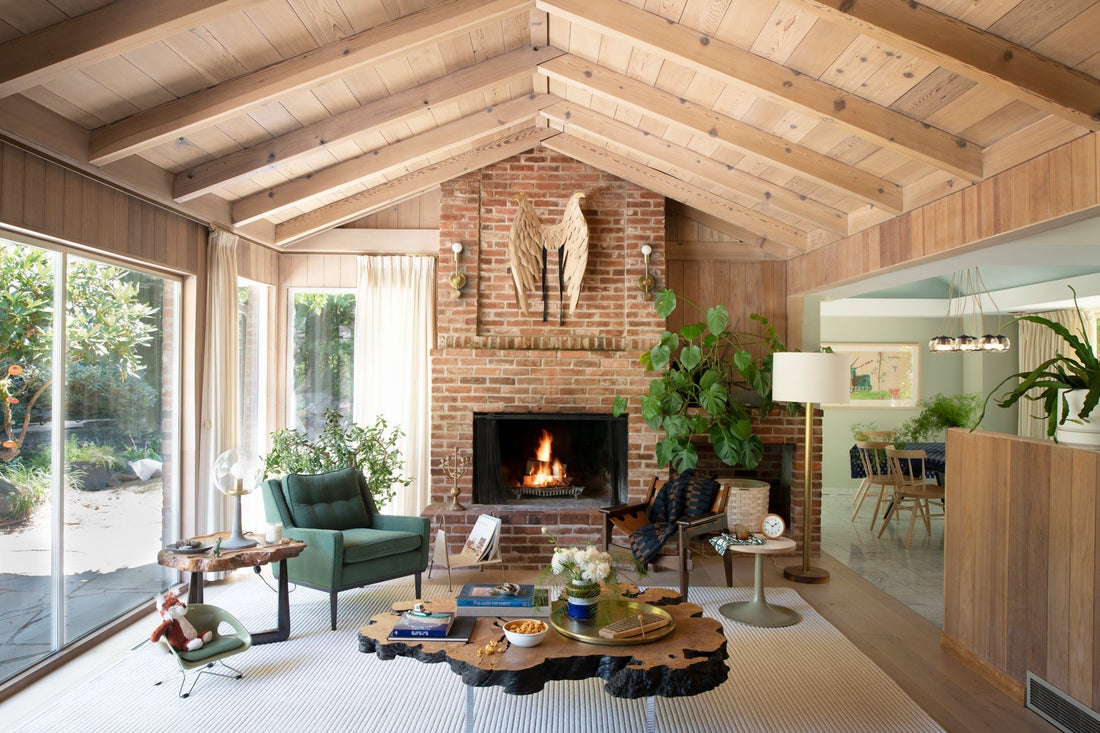 Living room with lit brick fireplace, vaulted wood ceiling, green armchair, coffee table, and sliding glass doors to a garden