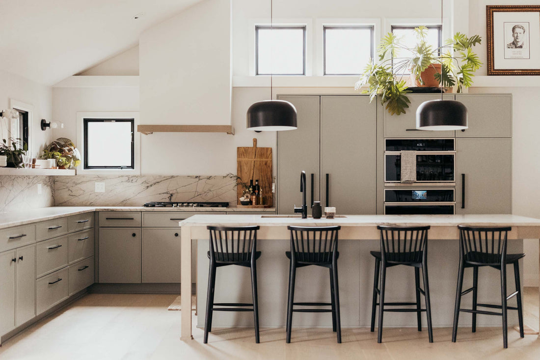 Open-plan kitchen with gray cabinets, marble backsplash, center island with four black bar stools and two black pendant lights, vaulted ceiling and potted plants