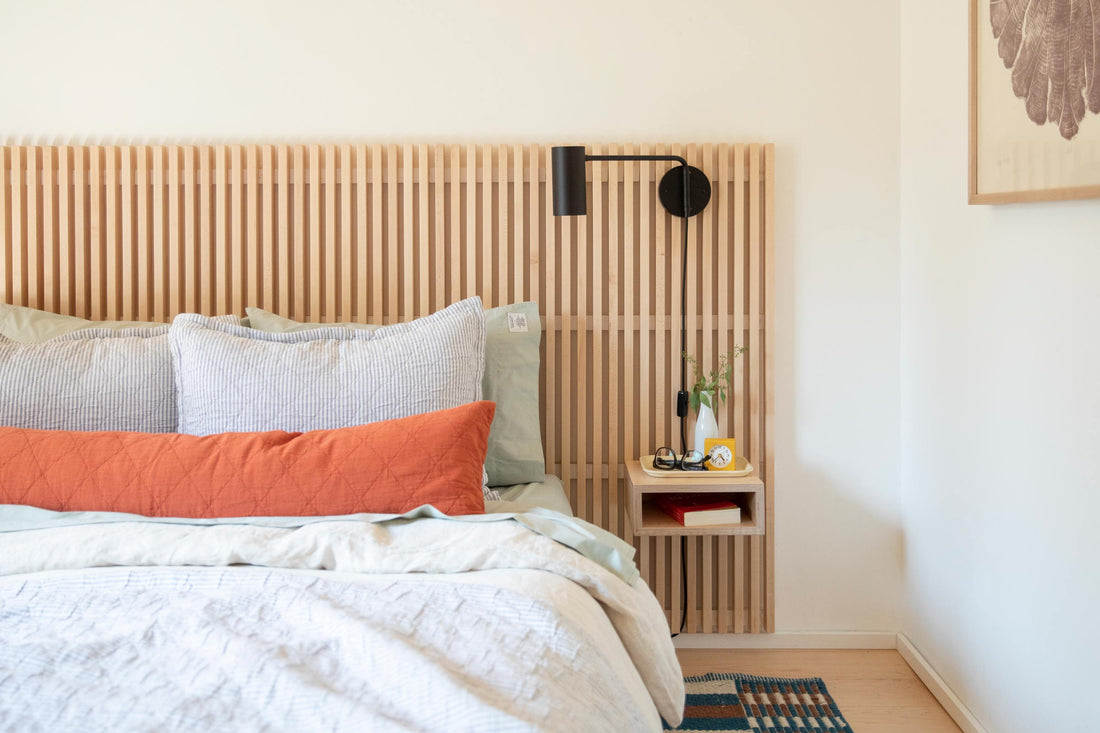 Bed with slatted wood headboard and black wall sconce beside a floating side table styled with a vase, yellow alarm clock, and glasses in a bright minimalist bedroom