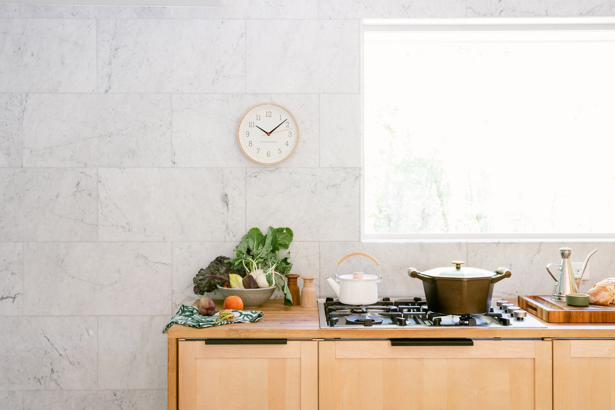 Cast-iron pot simmering on a gas stove in a bright marble-walled kitchen with a kettle, bowl of fresh vegetables, and a wall clock