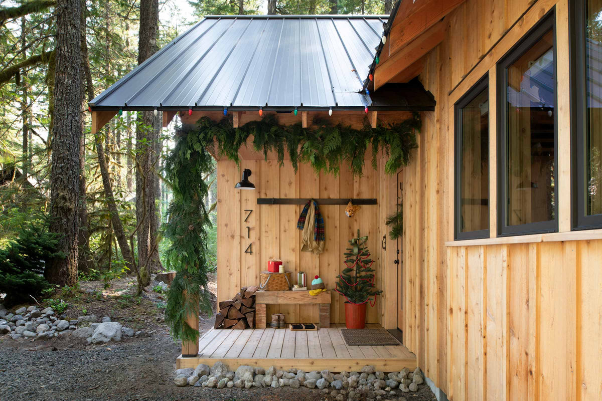 Cabin porch decorated with evergreen garland, colored string lights, a small potted Christmas tree, and stacked firewood