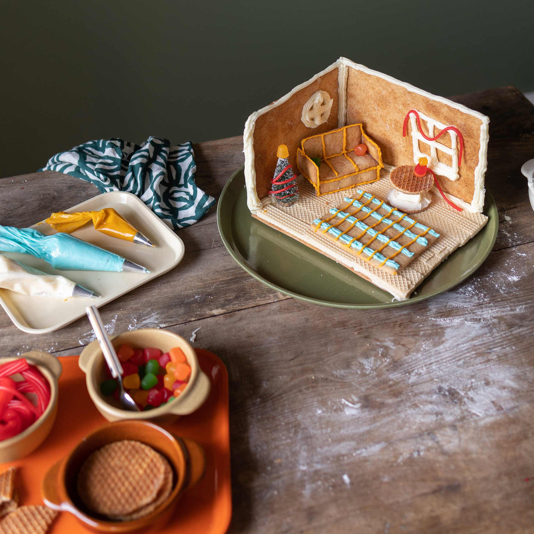 Mini gingerbread living room on a green plate with cookie sofa, wafer rug, iced window, tiny tree, and piping bags on a table