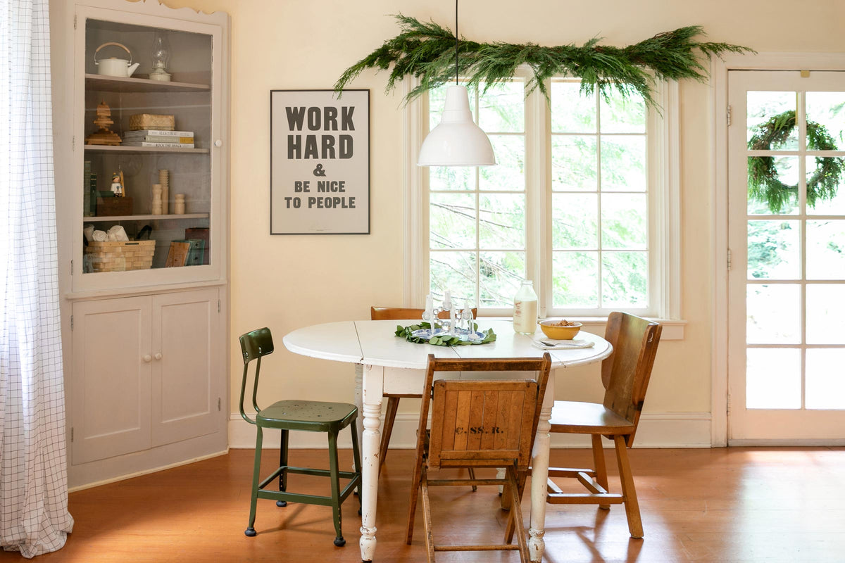 Dining nook with white table, vintage chairs, pendant light, greenery garland over the window and framed sign reading “Work Hard and Be Nice to People”