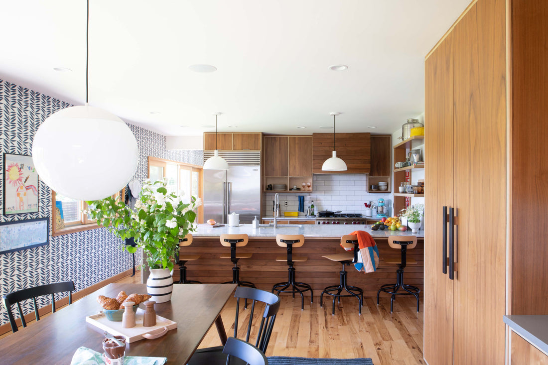 Sunlit kitchen and dining area with walnut cabinets, marble island, bar stools, patterned wallpaper, and vase of flowers