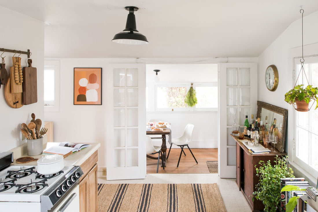 Sunlit tiny kitchen opening to a cozy dining nook with white chairs, wooden accents, hanging plants, striped rug, and sideboard with bottles