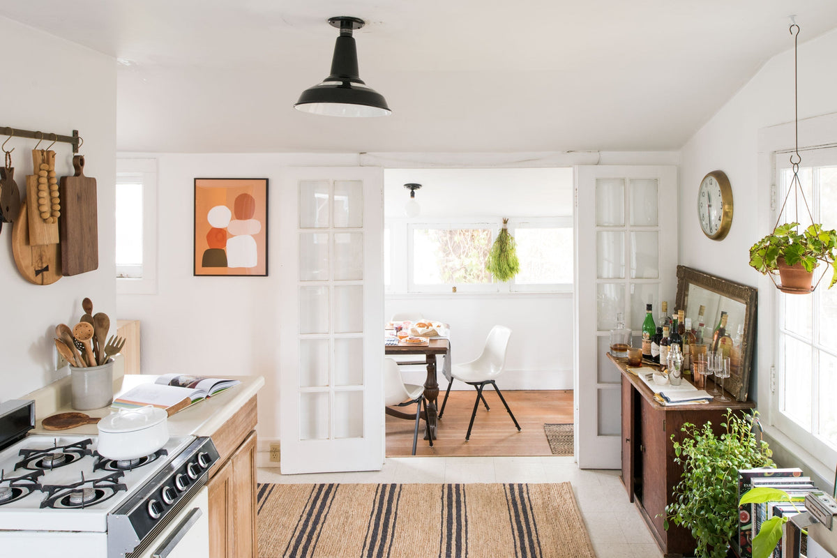 Sunlit tiny kitchen opening to a cozy dining nook with white chairs, wooden accents, hanging plants, striped rug, and sideboard with bottles