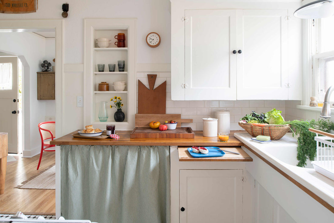 Sunlit traditional kitchen with white cabinets, wood countertops, an apron sink by a window and a basket of fresh produce on the counter