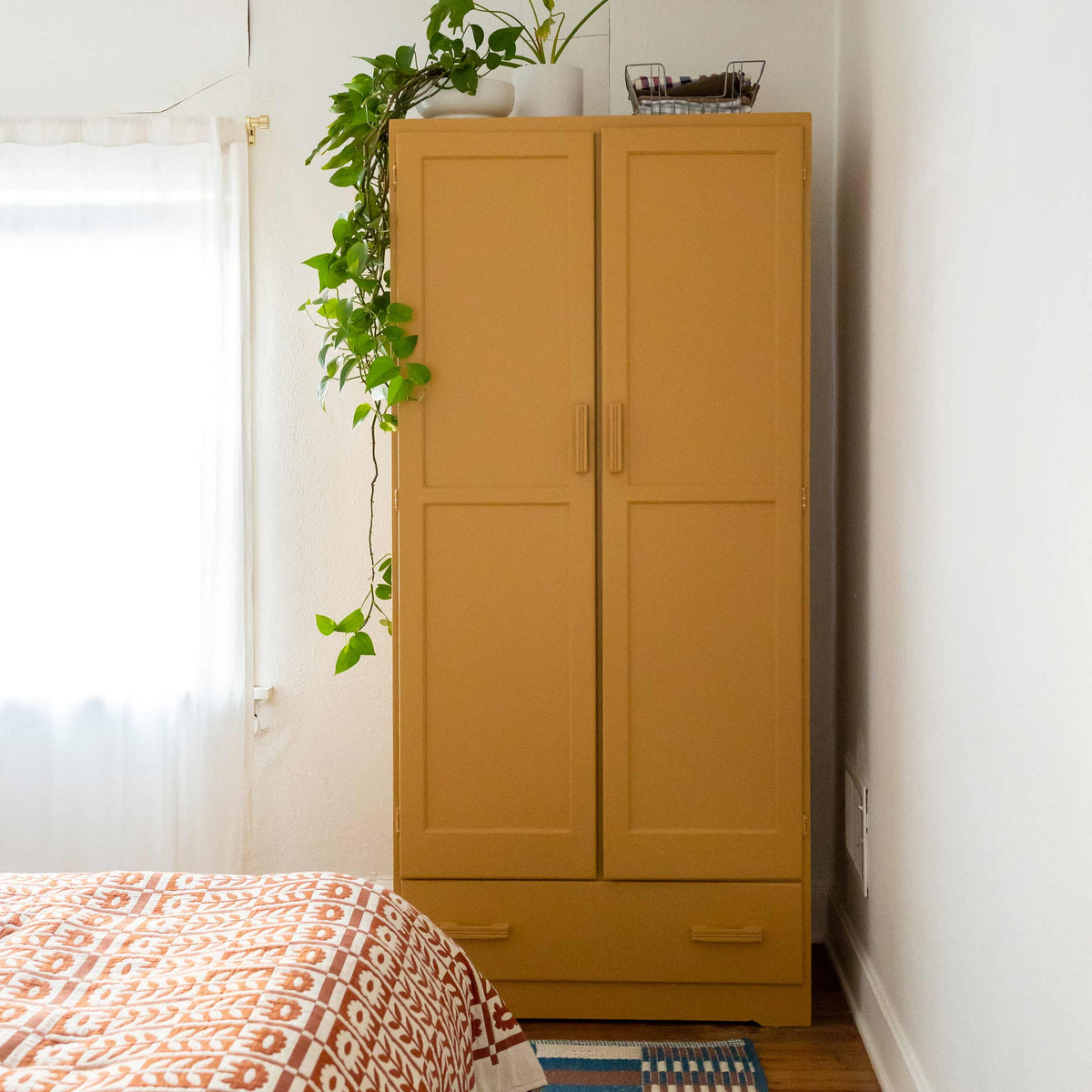 Front view of mustard armoire showcasing paneled doors and drawer pulls with trailing pothos and sheer window in bedroom