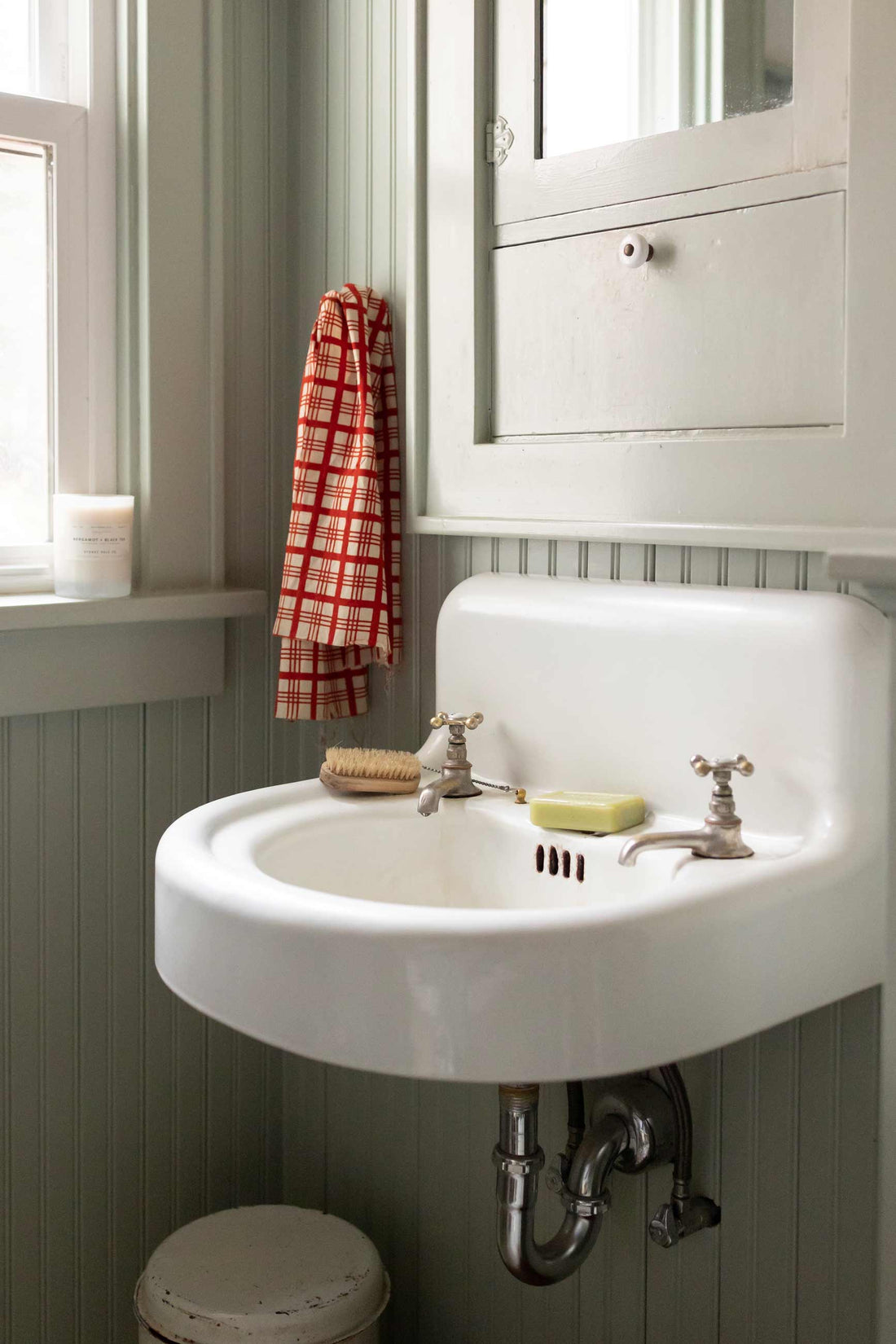 White vintage wall-mounted sink with brass taps, red checked hand towel on beadboard wall and a candle on the windowsill