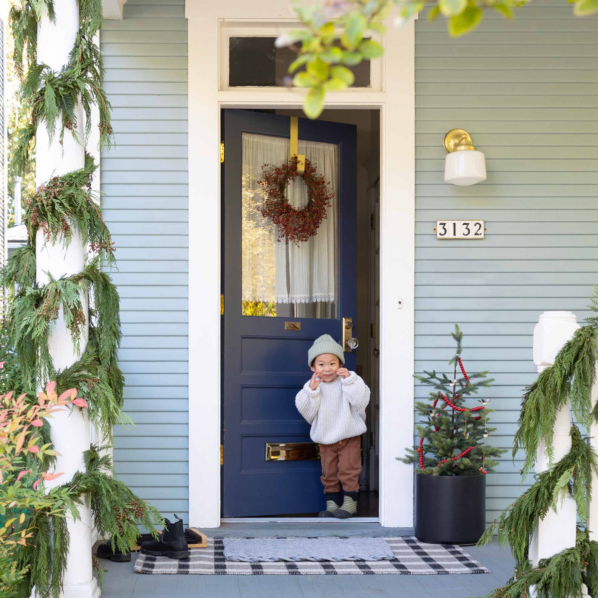 Toddler at a navy front door smiling on a porch with evergreen garlands, wreath, small decorated tree, house number 3132