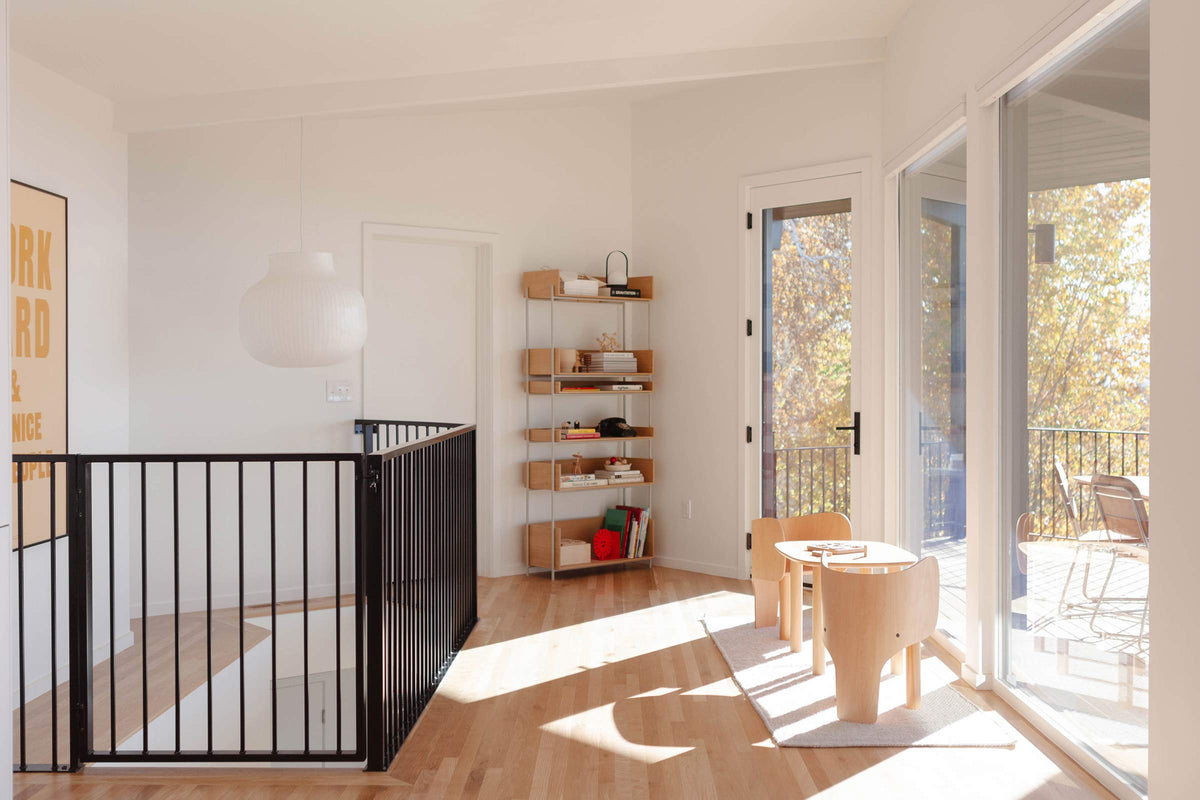 Sunlit kids’ play area with small wooden table and two chairs beside sliding glass doors, black stair gate, and a bookshelf