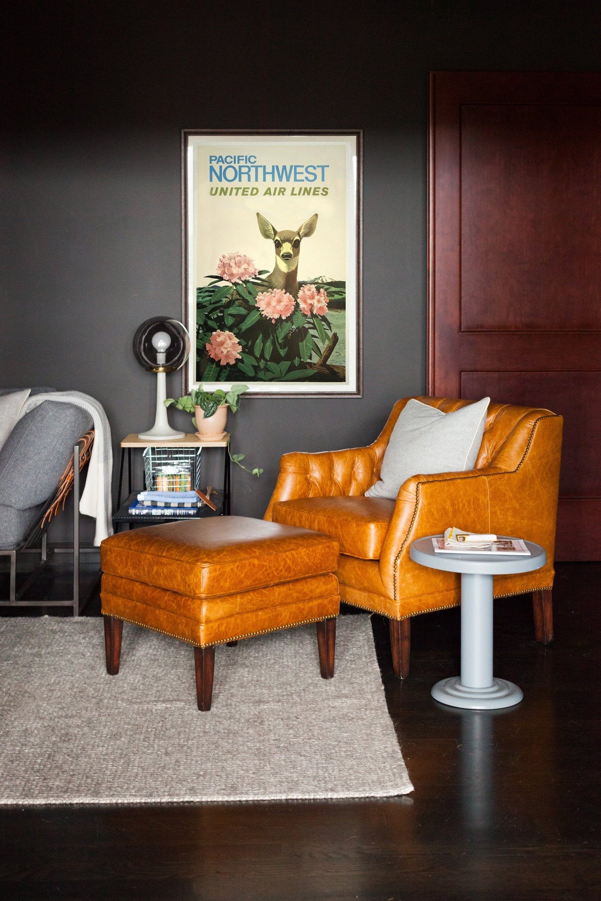 Leather cognac armchair with matching ottoman beside framed travel poster reading “PACIFIC NORTHWEST UNITED AIR LINES” on a dark gray wall