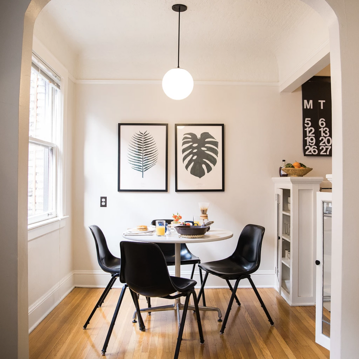 Luna globe pendant hanging above a round breakfast table in a bright dining nook with black chairs and framed botanical prints