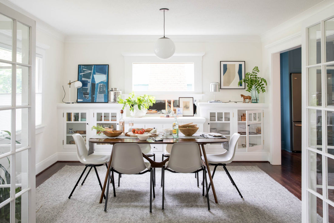 Dining room with wood table set for a casual meal, six white chairs, a globe pendant light, built-in white cabinets and potted plants