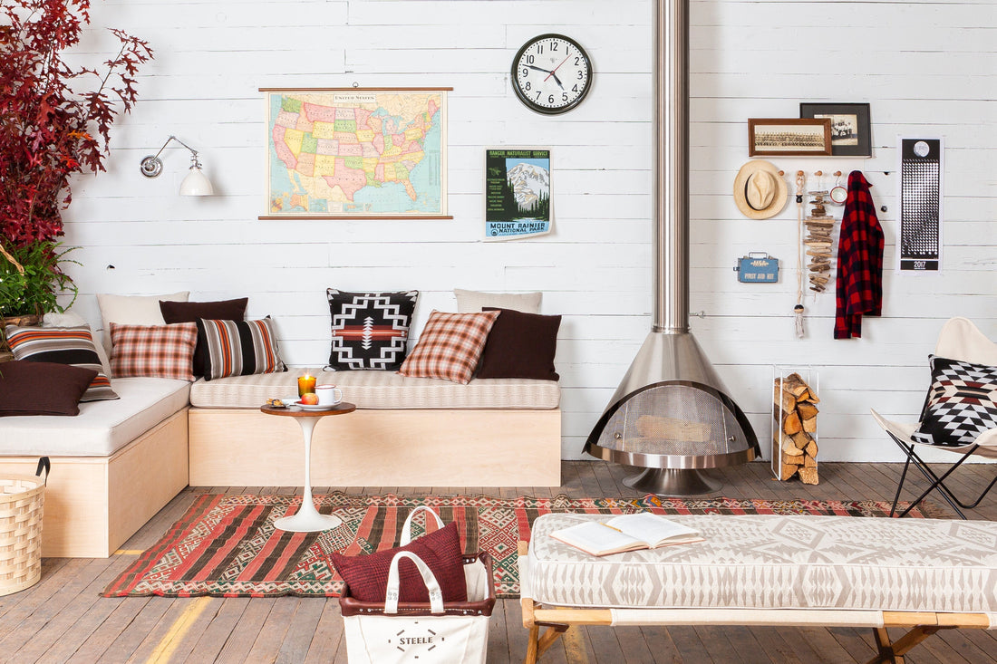 Living room vignette with bench seating, pillows, wall map and clock, freestanding metal fireplace and stacked firewood