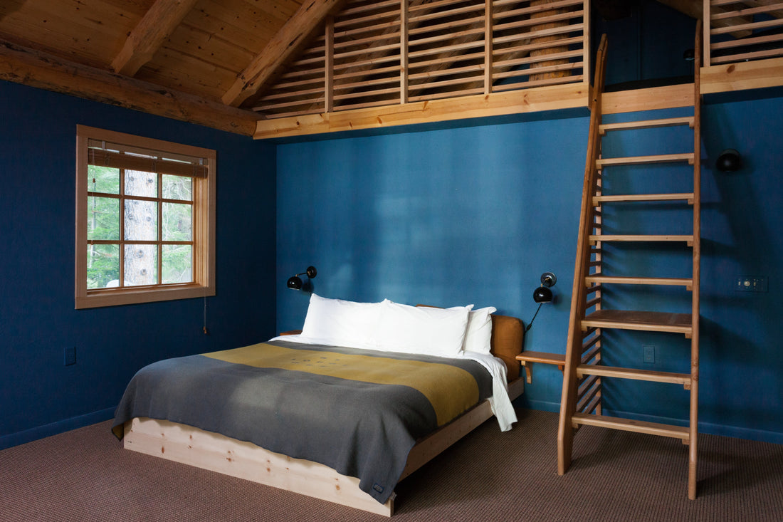 Cozy bedroom with low wood platform bed, white pillows, blue painted walls and a wooden ladder leading to a loft; window showing trees outside
