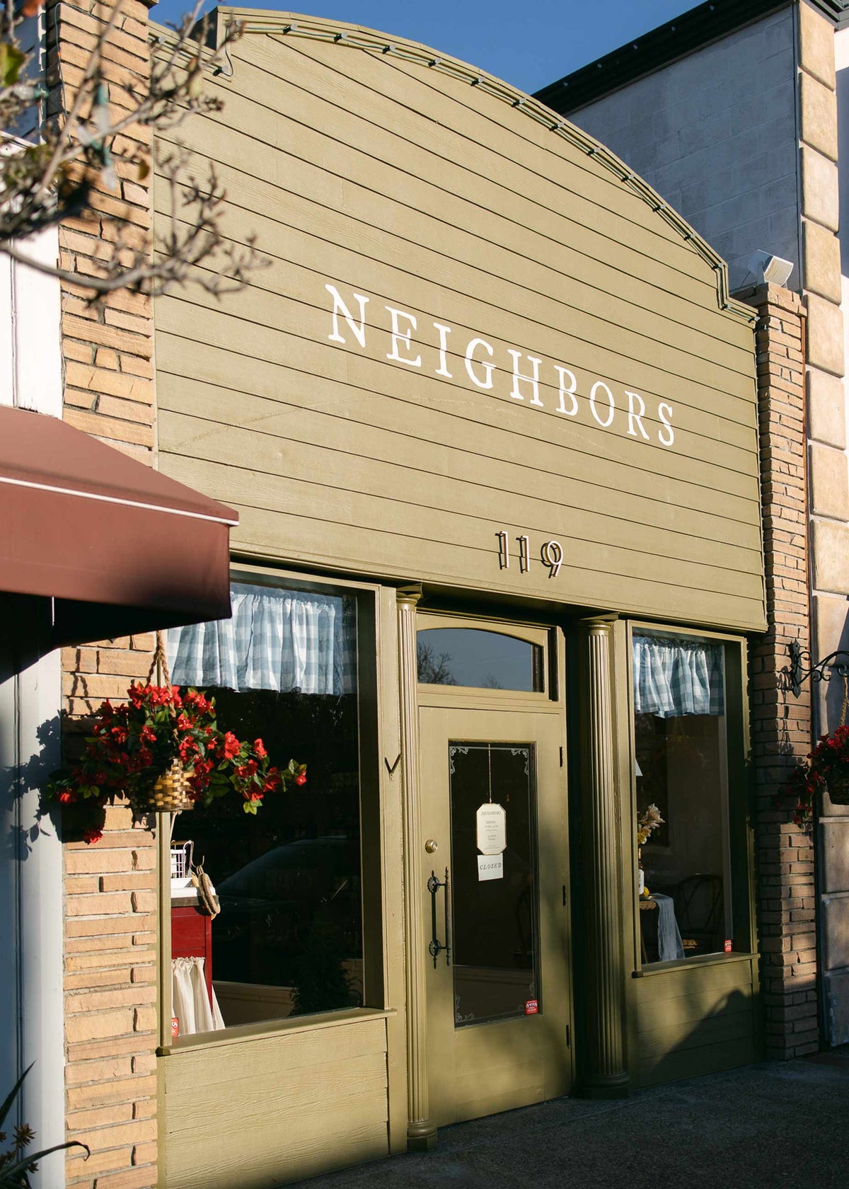 Neighbors general store storefront showing olive-green wood facade, white NEIGHBORS sign, hanging red flowers, and number 119