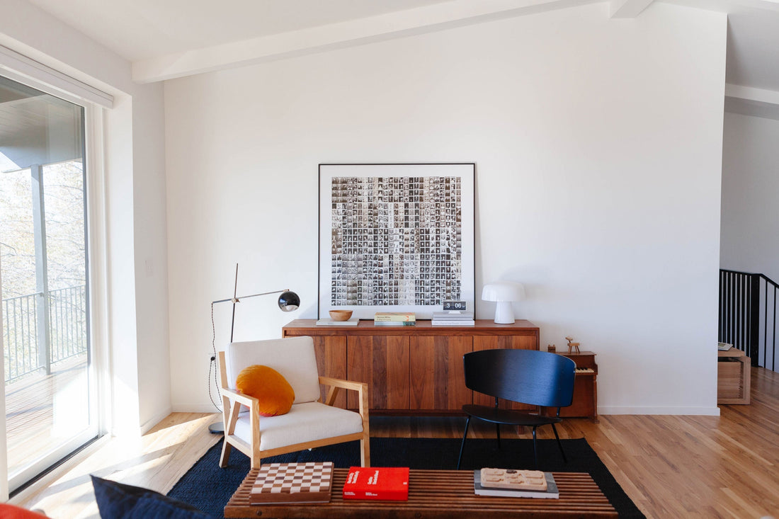 Sunlit minimalist living room with walnut sideboard, framed photo collage, armchair with orange pillow, and black accent chair