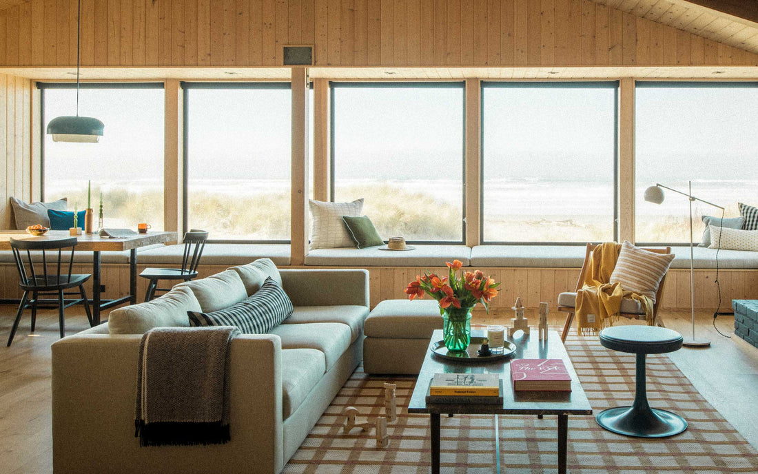 Sunlit living room with beige sectional and window seat facing sand dunes and the ocean through large picture windows