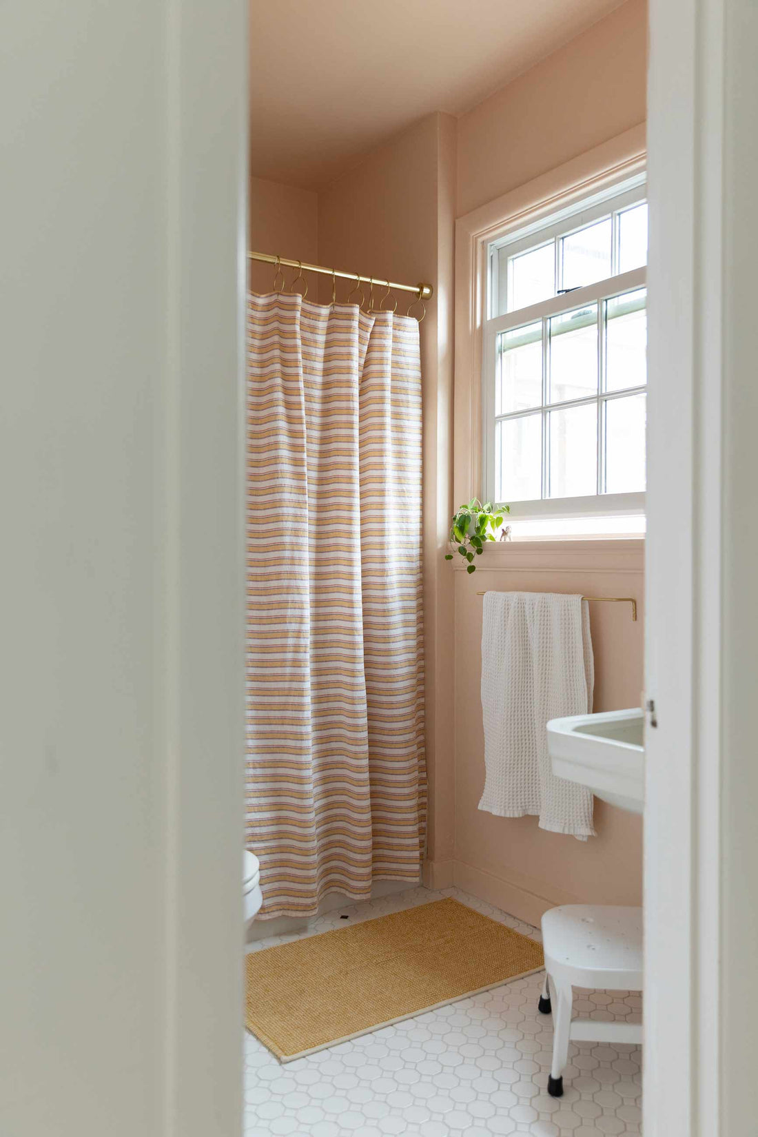 Small blush-pink bathroom seen through a doorway with a striped shower curtain on a brass rod, pedestal sink, towel on a gold rail, and a window plant
