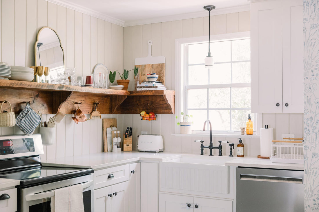 Sunlit farmhouse kitchen with white cabinets, apron-front sink and dark faucet, reclaimed wood open shelf holding dishes and a potted plant