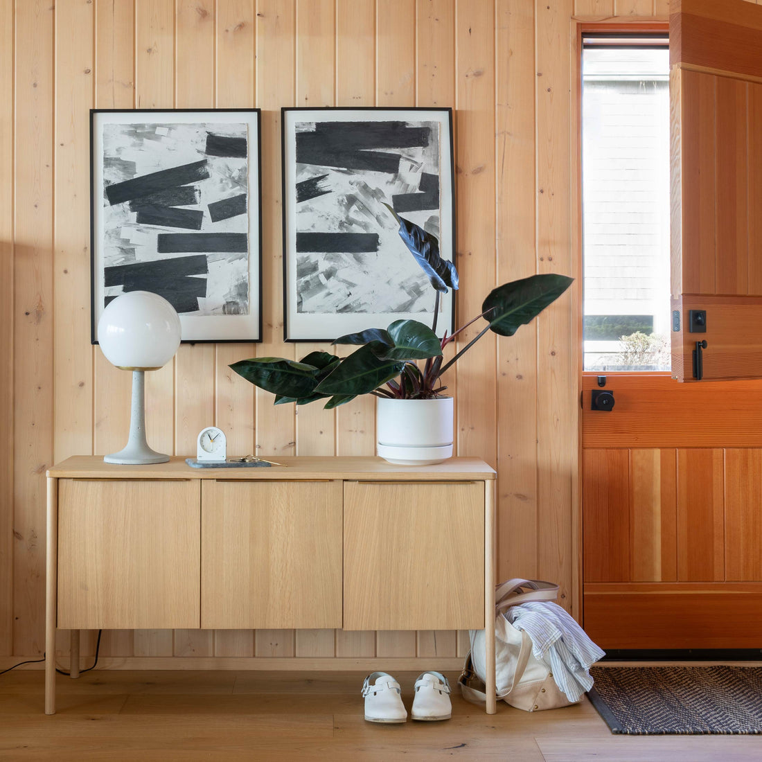 Sideboard with globe lamp, clock, potted dark-leaf plant, and two abstract black-and-white prints in a wood-paneled entryway