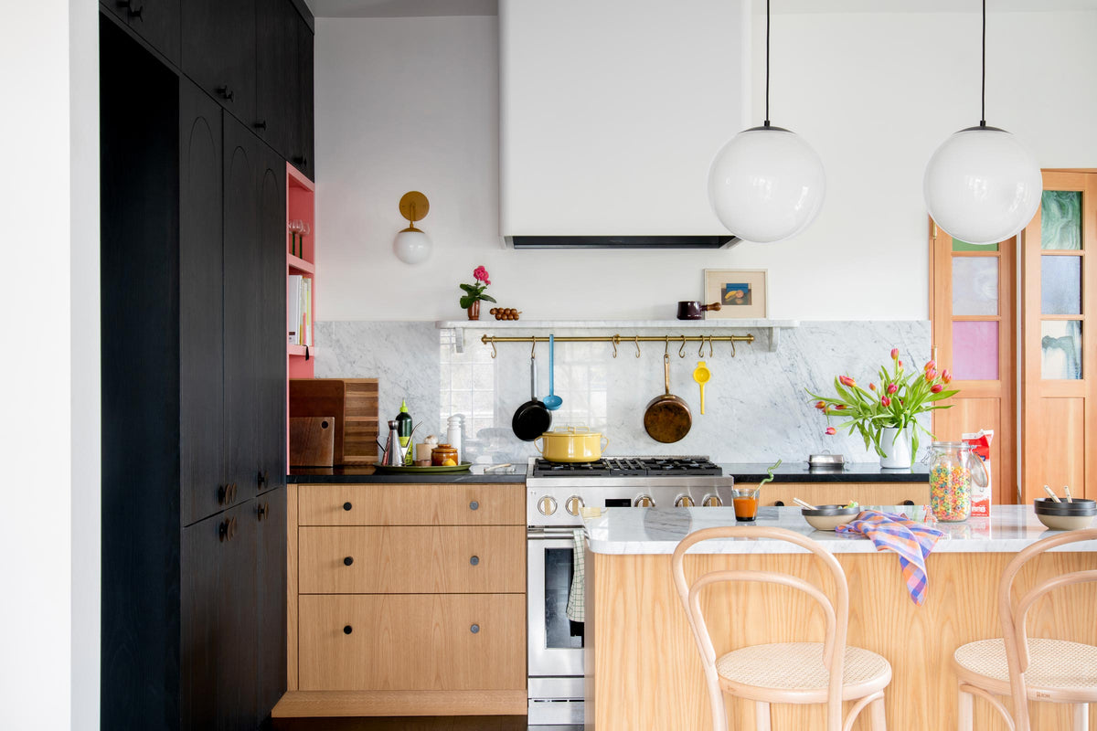 Bright modern kitchen with two white globe pendant lights over a marble island, light wood cabinets, stainless gas range, brass rail with hanging pans, and a vase of tulips on the counter