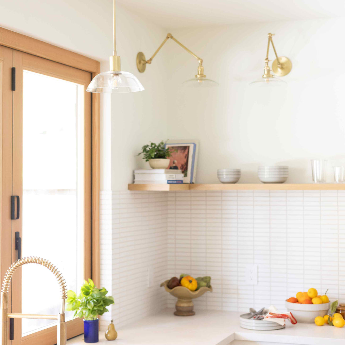 Clear glass pendant and brass swing arm sconces in a bright kitchen with white tile backsplash, wood shelf, blue potted herb, and fruit bowls